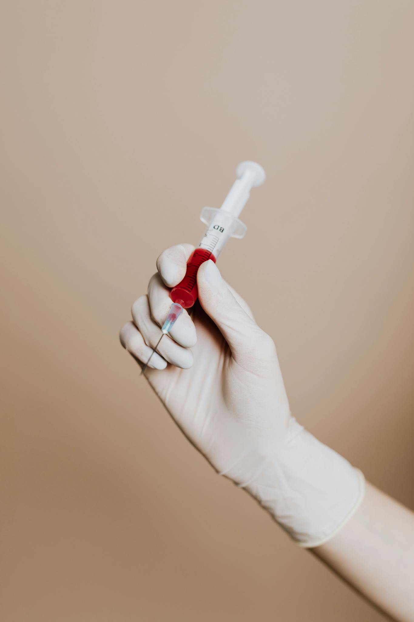A gloved hand holds a syringe filled with red liquid, set against a neutral background.